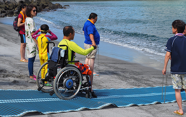砂浜に敷いたモビマットの上を車いすが自力で波打ち際に進む様子